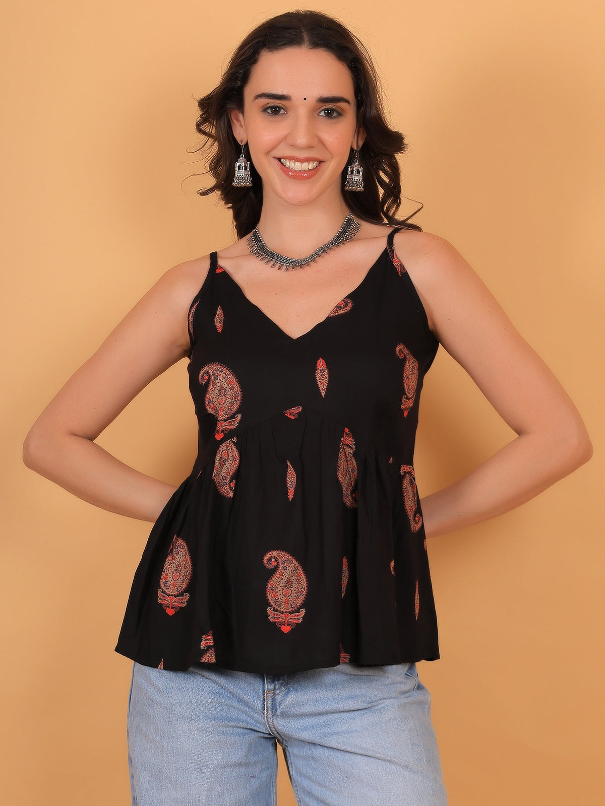 A female model wearing the ZUACC Indira Fit & Flare Ethnic Top, featuring a sleeveless relaxed-fit peplum silhouette in black with a golden traditional motif print, styled with blue denim jeans.
