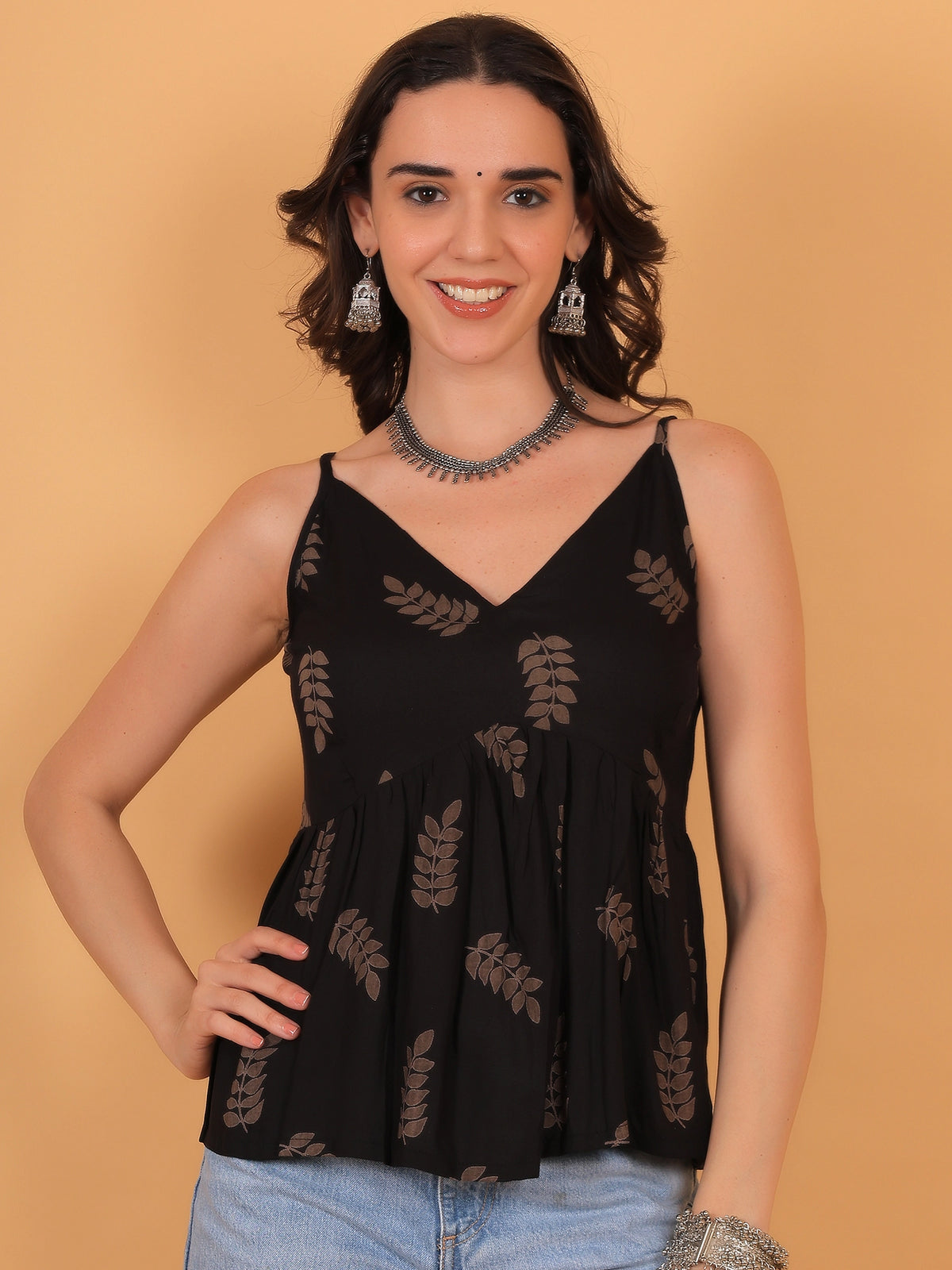 A female model wearing the ZUACC Vanya fit and flare ethnic top, featuring a sleeveless V-neck peplum silhouette in black with a brown leaf print pattern, styled with light blue jeans.