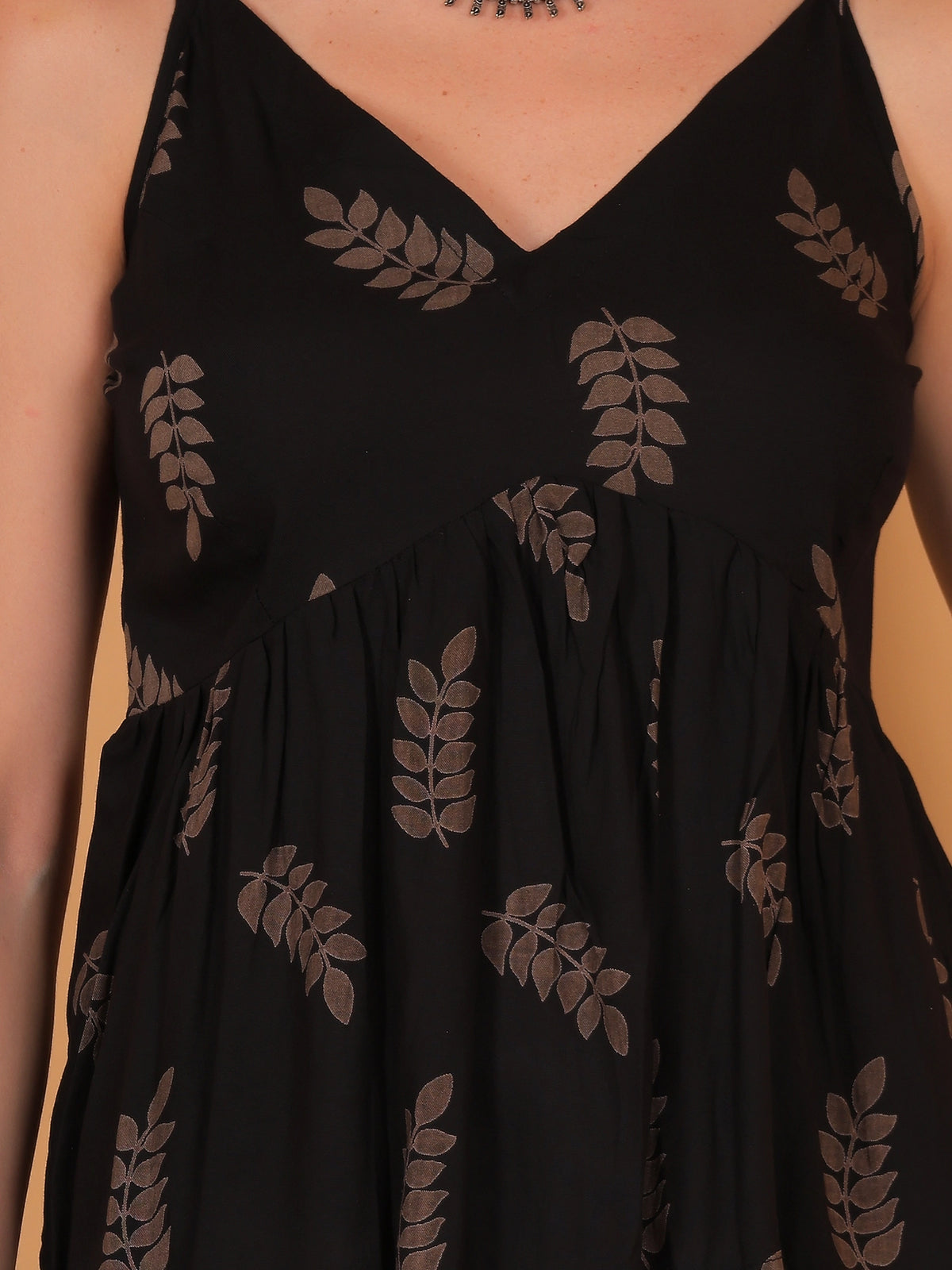 Front close-up shot of the Vanya black sleeveless top, showcasing the detailed brown leaf motif print on opaque rayon fabric against a warm peach-beige background.
